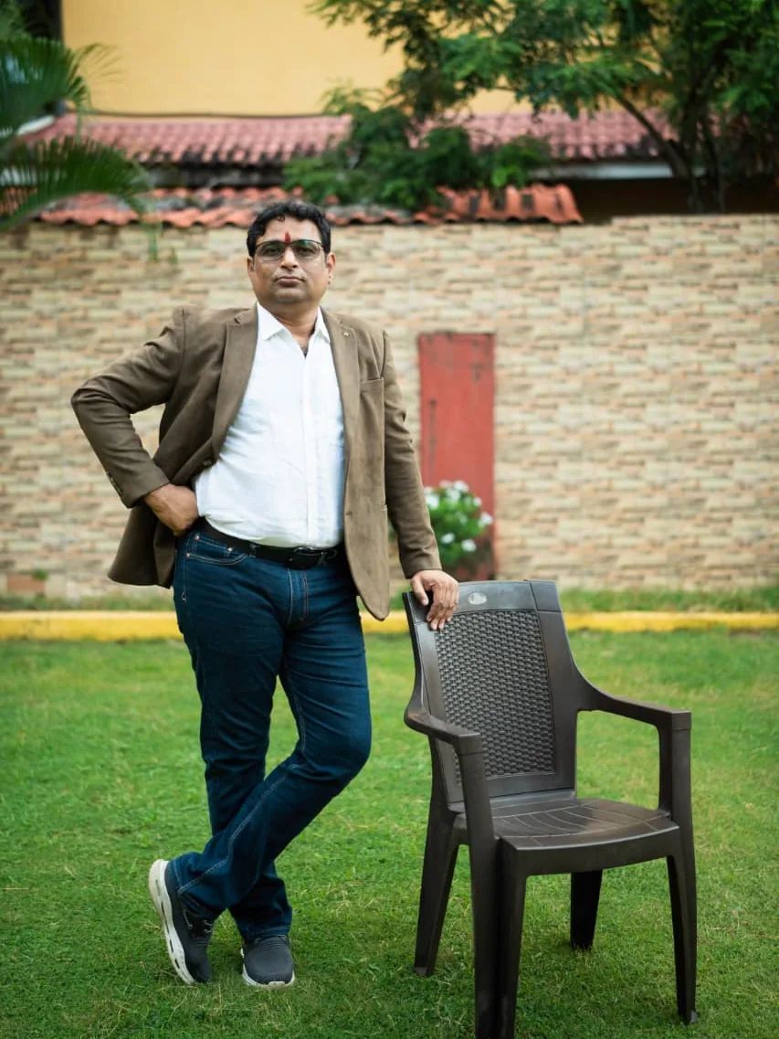 Prakash Shastry in a brown blazer, standing on a lawn beside a chair