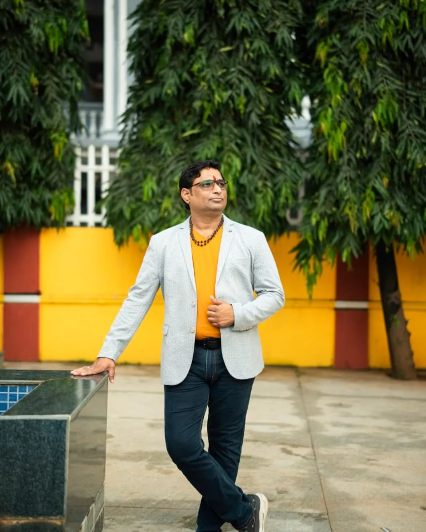 Prakash Shastry in a light grey blazer, leaning on a poolside wall in front of tall trees