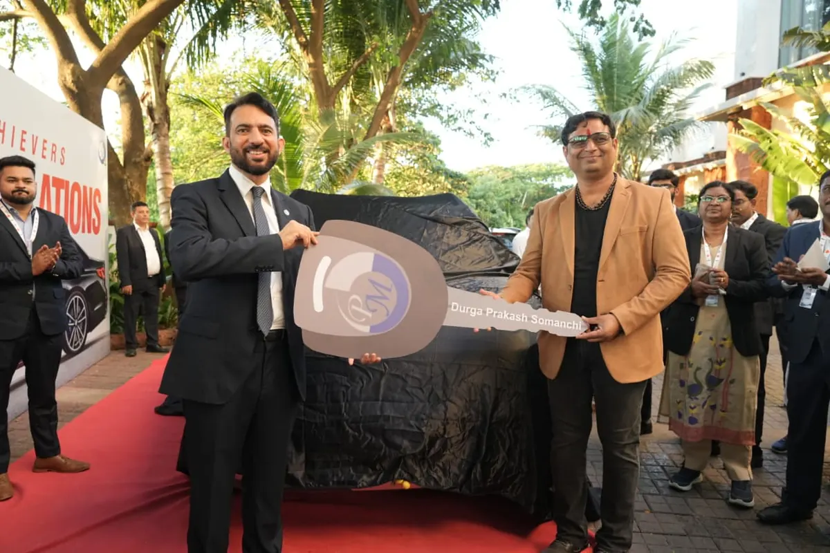 Prakash Shastry Somanchi receiving a PM International key bearing his name from a company representative at an achievers event, a covered car behind them