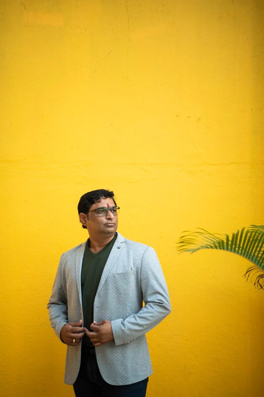Prakash Shastry Somanchi in a grey blazer, looking into the distance against a bright yellow wall
