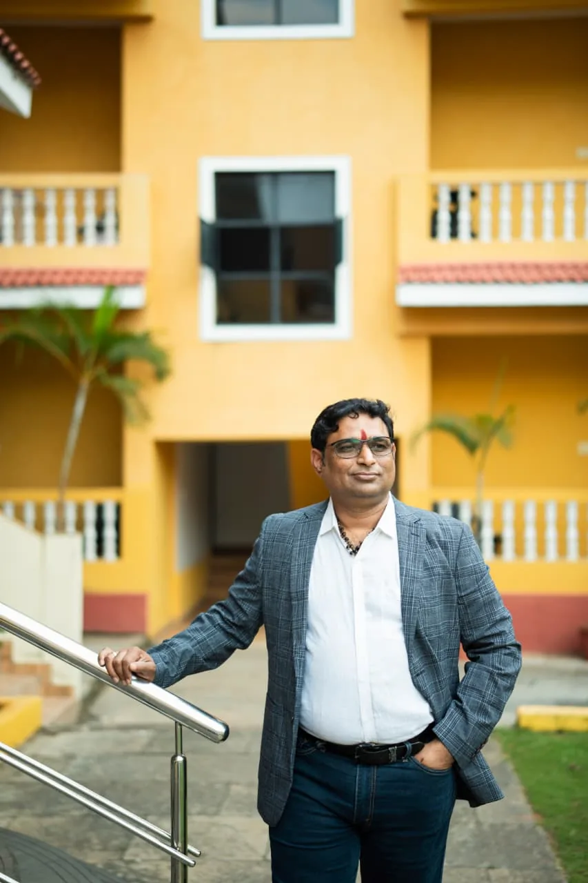 Prakash Shastry Somanchi smiling, resting one hand on a railing in a grey-blue blazer