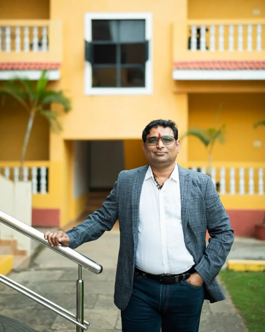 Prakash Shastry in a grey checked blazer, one hand on a stair railing, outside a yellow building