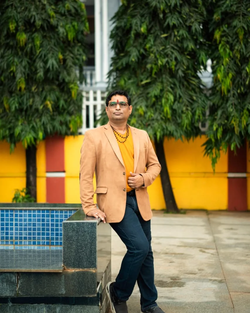 Prakash Shastry in a tan blazer, leaning on a poolside wall in front of tall trees