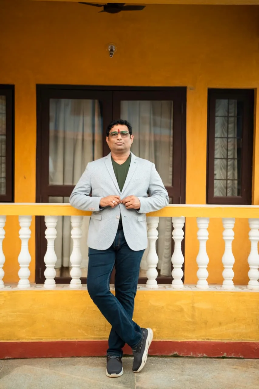 Prakash Shastry Somanchi leaning on a yellow balcony railing, hand on his blazer button